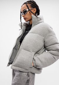 Kvinde med fletninger iført grå pufferjakke, sort skjorte, grå jeans og sølvfarvede solbriller, poserer mod hvid baggrund.