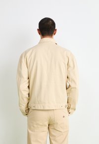 Beige bomullsjakke med et enkelt design, som har krage, lange ermer og knappelukking ved ermene. Viser en glatt tekstur og praktiske detaljer.