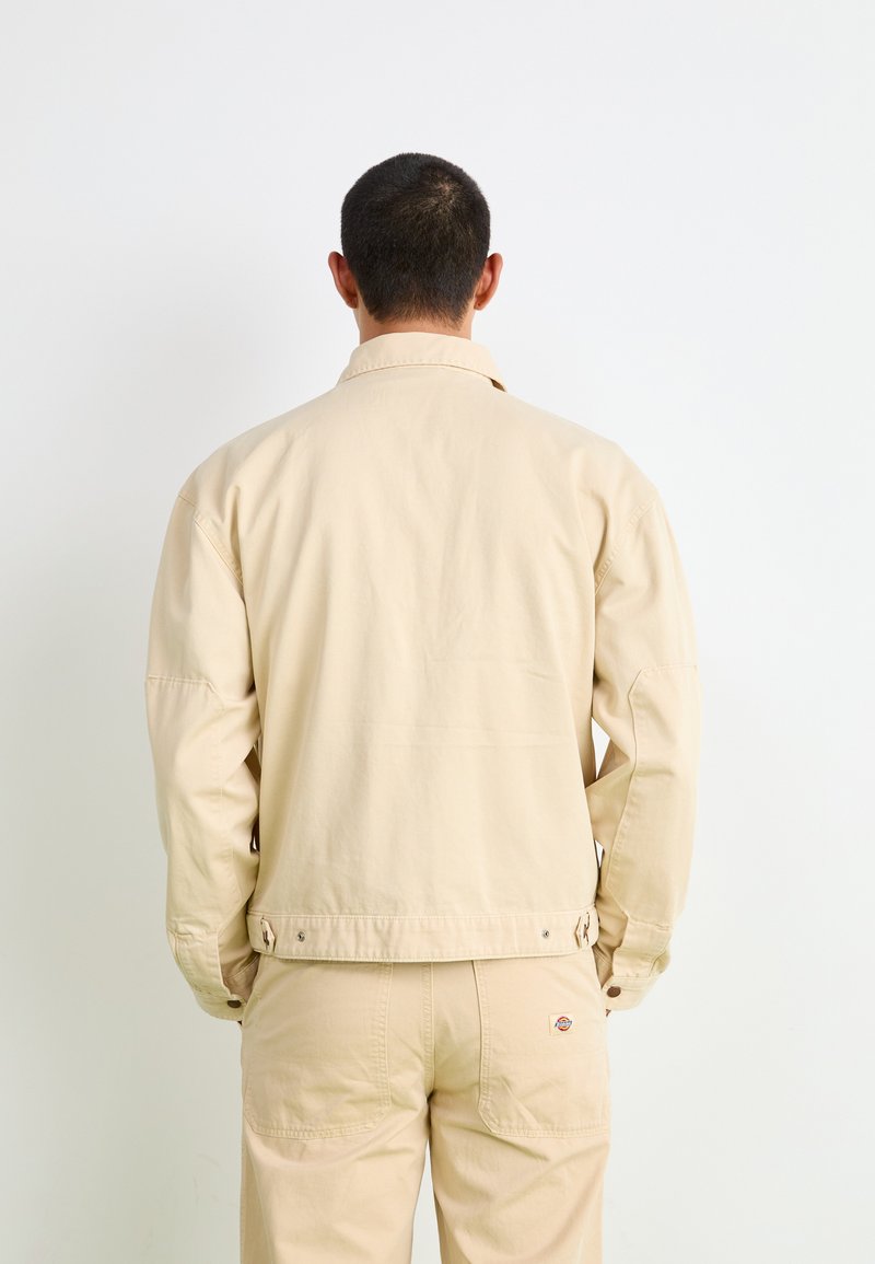 Veste en coton beige au design simple, dotée d'un col, de manches longues et de poignets boutonnés. Présentant une texture lisse et des accents pratiques.
