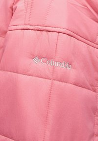 Ροζ τμήμα υφάσματος με καπιτονέ ύφασμα ενός μπουφάν με λευκό κεντημένο λογότυπο Columbia και λεπτομέρειες ραφής.