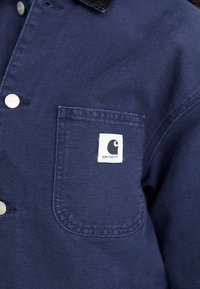 Primo piano di una giacca Carhartt blu navy che mostra la tasca frontale con il patch del logo bianco e bottoni a scatto argentati.