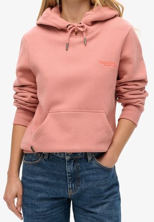 Person trägt einen rosa SuperDry Hoodie mit Fronttasche, Kordelzugkapuze und blauen Jeans, steht mit einer Hand in der Tasche.