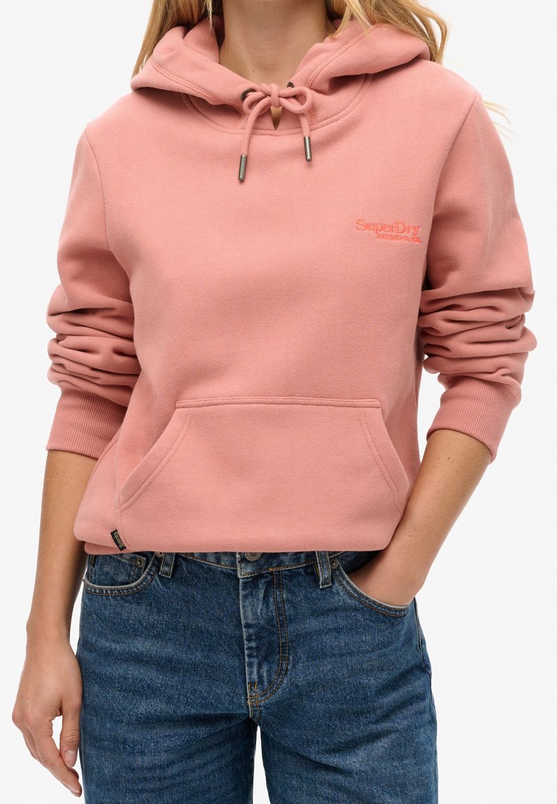 Personne portant un sweat à capuche rose SuperDry avec une poche avant, une capuche à cordon et un jean bleu, debout avec une main dans la poche.