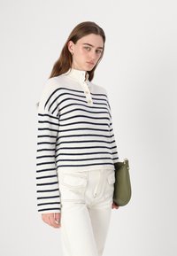 Stripet strikkegenser i kremfarge og marineblå med høy krage og tre knapper. Sammensatt med hvite bukser og en grønn clutchtaske.