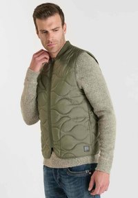 Gequilte groene vest met een ritssluiting, voorzien van gestructureerde golvende patronen. Gecombineerd met een lichtgrijs sweater en blauwe jeans.