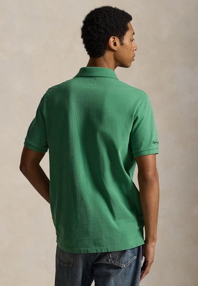 Man met kort krullend haar, gekleed in een groen poloshirt en een blauwe spijkerbroek, met zijn rug naar de camera en tegen een neutrale achtergrond.