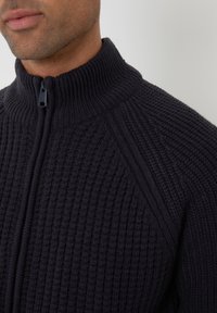 Dunkel gestrickter Pullover mit hohem Kragen und halbem Reißverschluss. Verfügt über eine gerippte Textur und winkelförmige Schulternaht für eine verbesserte Passform.