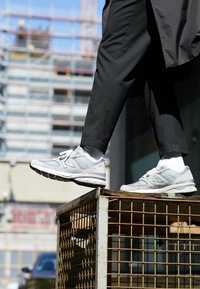 Personne portant un pantalon noir et des baskets grises avec des chaussettes blanches marchant sur une cage en métal rouillé en plein air dans un cadre urbain.