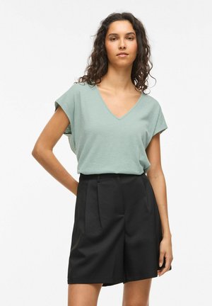 Woman with wavy hair wearing a light green V-neck short sleeve top and high-waisted black shorts, standing with one hand behind her back.