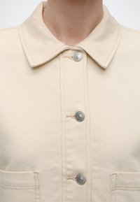 Κοντινό πλάνο του μπροστινού μέρους μπεζ τζάκετ με μεταλλικά κουμπιά με πατητά κουμπώματα και μυτερό γιακά, που δείχνει το ανώτερο μέρος του στήθους και της περιοχής του λαιμού.