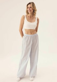 Biały krótki top na ramiączkach zestawiony z jasnoniebieskimi spodniami typu wide-leg w białą kratkę. Modelka stoi z rękami w kieszeniach, mając na sobie białe buty.