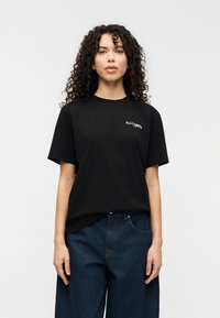 Μαύρο κοντο μάνικο t-shirt από βαμβάκι, με λογότυπο στο αριστερό στήθος. Συνδυασμένο με φαρδιά μπλε τζιν παντελόνια.