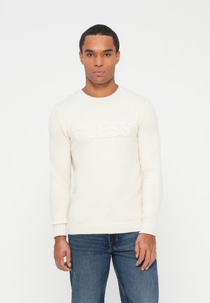 Suéter de punto color crema con cuello redondo, que presenta el texto elevado "GUESS" en el pecho. Lleva puestas jeans de mezclilla azules.