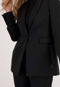 Vrouw draagt een strakke zwarte blazer en zwarte broek, houdt haar blonde haar met één hand bij haar gezicht.