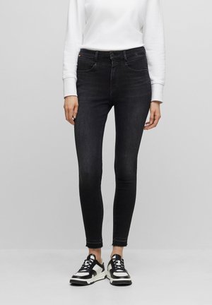 Femme portant un jean skinny noir aux ourlets effilochés, une chemise blanche à manches longues et des baskets noires et blanches, debout devant un fond uni.
