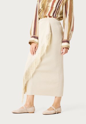 Mujer con falda midi acanalada color crema con detalle de flecos diagonales, blusa de rayas de manga larga y zapatos planos beige con correa y hebilla.