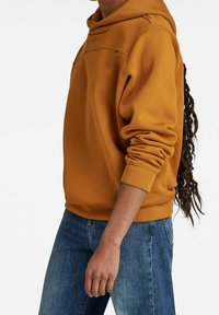 Sudadera amarilla mostaza con un bolsillo tipo canguro, mangas con puños y un corte holgado, combinada con jeans de mezclilla azul. Tela texturizada visible.