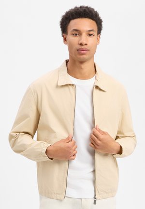 Jonge man draagt een beige jack met rits over een wit T-shirt, staande tegen een witte achtergrond, en houdt de zijkanten van het jack bij de ritssluiting vast.
