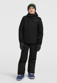 Zwarte sneeuwjas met een capuchon, ritszakken en paarse handschoenen. Zwarte sneeuwbroeken en blauwe laarzen met witte veters maken de outfit compleet.