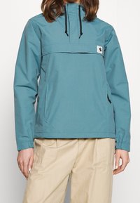 Vrouw in een teal anorakjack met zwarte trekkoorden en beige broek, staand tegen een effen witte achtergrond.