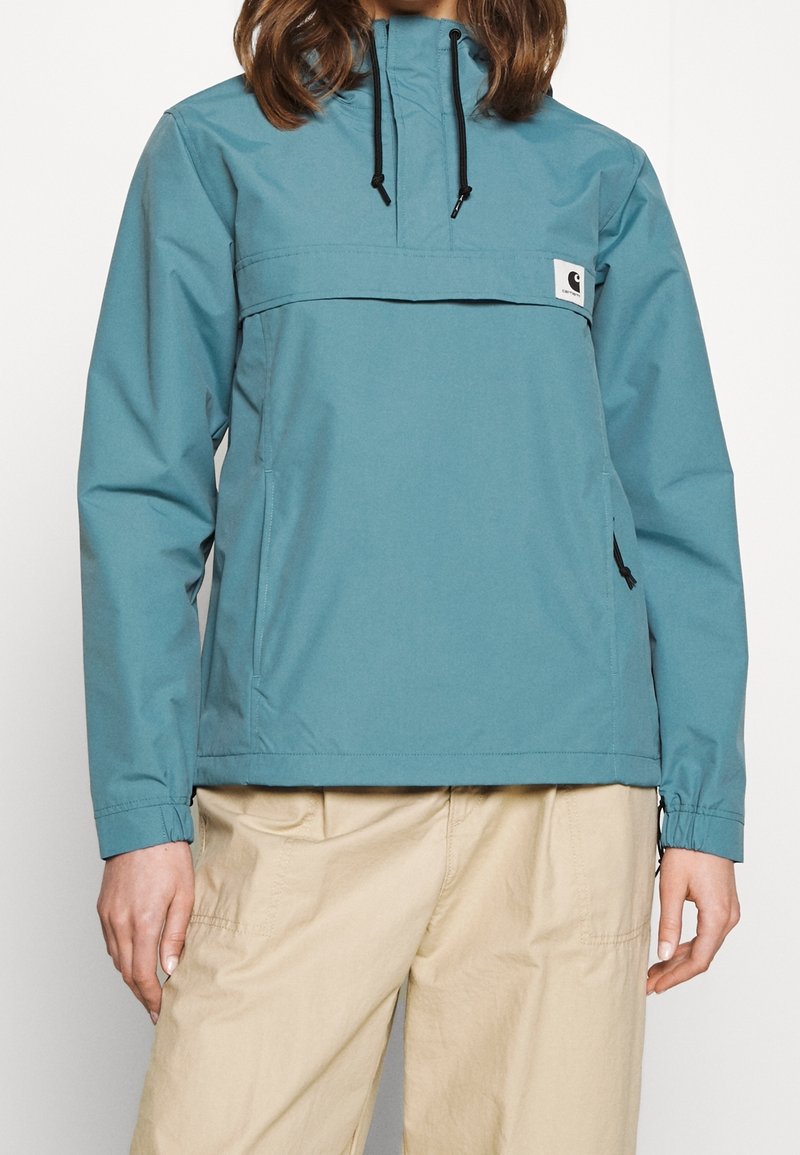 Vrouw in een teal anorakjack met zwarte trekkoorden en beige broek, staand tegen een effen witte achtergrond.