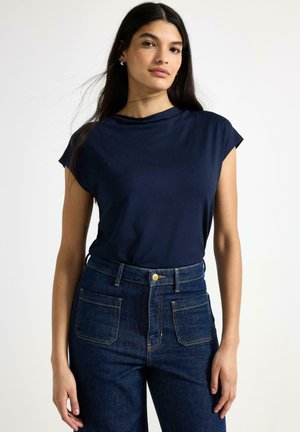 Ung kvinde med langt mørkt hår bærer en marineblå top med korte ærmer og højtaljede mørkeblå jeans mod en hvid baggrund.