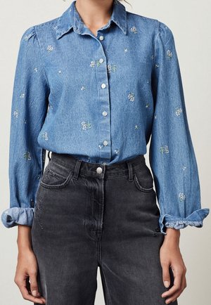 Blue denim button-up shirt with small floral embroidery, rolled cuffs, paired with high-waisted faded black jeans.