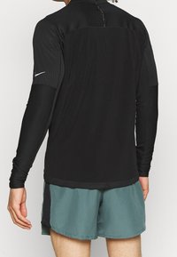 Persoon in een zwart sportshirt met lange mouwen en geribbelde mouwen, met groene geperforeerde sportshorts, gezien vanaf de achterkant.