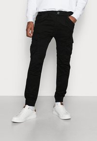 Pantalones cargo negros con puños elásticos, que cuentan con bolsillos laterales y cierre de botón. Combinados con zapatillas blancas y una sudadera blanca.