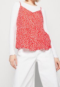 Top rojo sin mangas con tirantes de espagueti y lunar blanco, con botones en la parte frontal y cintura fruncida, usado sobre una camisa blanca de manga larga.