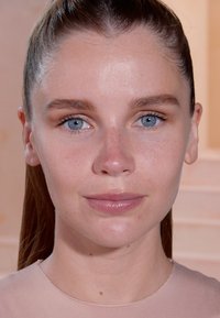 Vrouwelijk model met steil bruin haar in een paardenstaart, een egale huid, lichte make-up, en gekleed in een aansluitende, huidkleurige top met een gladde textuur.