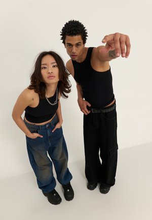 Two individuals stand in casual attire: one in a black tank top and oversized blue jeans, the other in a black tank top and black cargo pants.