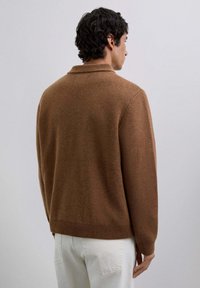 Mand med sort krøllet hår iført en brun strikket sweater og hvide bukser, stående mod en ensfarvet lys grå baggrund, vendt væk.