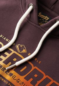 Bordowy bawełniany hoodie z białymi sznurkami ściągającymi, z logo o pomarańczowo-żółtej gradientowej strukturze i metalowym detalem.