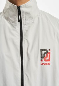 Light grey windbreaker jacket with a high collar, front zipper, and a prominent black and red graphic logo on the left chest.