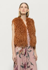 Braune, struppige Kunstpelzweste mit offener Vorderseite und geschichtetem Textur, kombiniert mit einem rosa floral gemusterten Kleid. Ärmelloses Design.