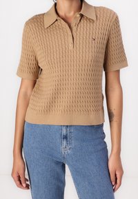 Osoba w beżowym polu z krótkim rękawem w splot warkoczowym i niebieskich jeansach z wysokim stanem, z czarnym kwiatowym tatuażem na prawej przedramieniu.