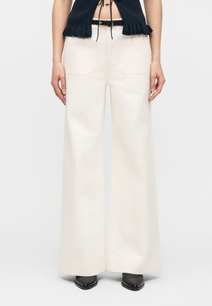 Kobieta ubrana w kremowe spodnie o wysokim stanie i szerokich nogawkach z przednimi naszywanymi kieszeniami, czarny pasek, czarny top bez rękawów oraz czarne spiczaste buty.