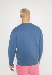 Persona con capelli corti biondi che indossa una felpa blu e pantaloni rosa, di spalle verso un muro bianco semplice.
