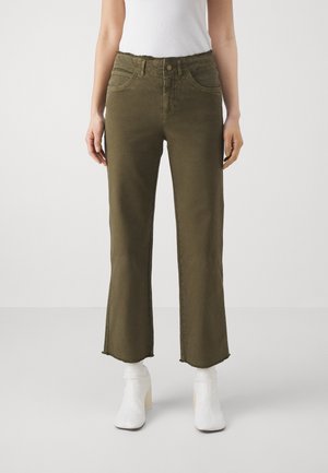 Mujer con pantalones anchos cortos de color verde oliva con bajos sin rematar y botas blancas hasta el tobillo, de pie sobre un fondo claro y liso.