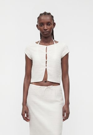 Femme aux cheveux tressés portant un crop top blanc à manches courtes boutonné et une jupe assortie à cordon sur fond uni.