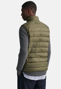 Chaleco acolchado de color verde oliva con diseño acolchado, cuello alto y material ligero, superpuesto sobre una camiseta de manga larga gris.