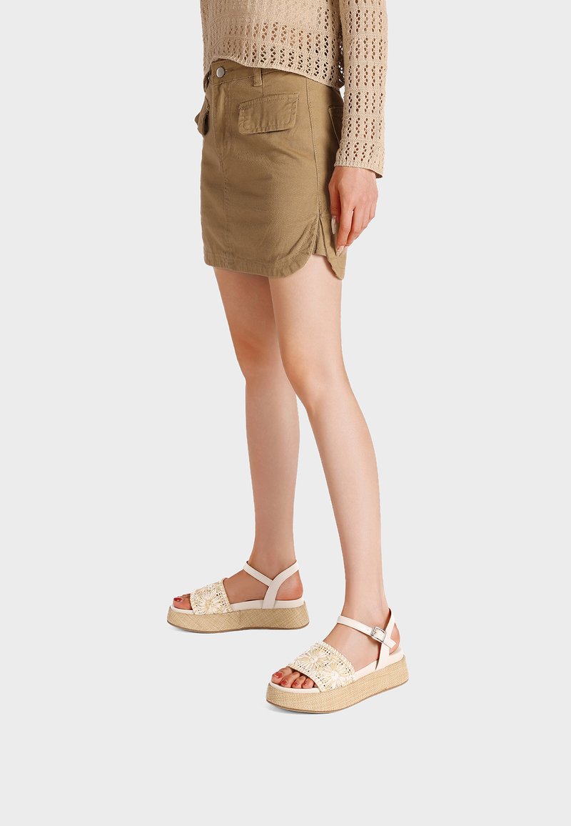 Mujer con suéter de ganchillo beige, minifalda marrón con abertura lateral y sandalias beige con plataforma y detalles florales, de pie sobre fondo blanco.