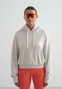 Persona con sudadera beige, leggings rojo-naranja, aretes de oro en forma de aro y grandes gafas de sol deportivas rojas y reflectantes, de pie contra un fondo liso.