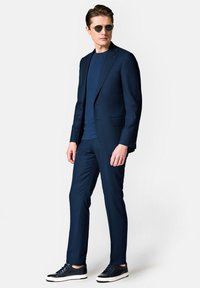 Granatowy garnitur wykonany na miarę z klapami w kształcie notcha, połączony z dopasowaną koszulką i ciemnymi sneakersami. Okulary przeciwsłoneczne w srebrnym odcieniu uzupełniają stylizację.