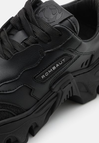 Schwarze Sneaker aus einer Mischung aus glattem und strukturiertem Leder, markanter Markenbezeichnung, dicker Sohle mit gezacktem Muster und schwarzen Schnürsenkeln.