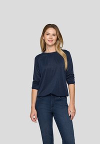 Junge Frau mit langen blonden Haaren, die ein navyblaues Langarmoberteil und dunkelblaue Jeans trägt, steht vor einem schlichten grauen Hintergrund.