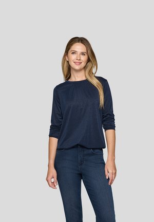 Junge Frau mit langen blonden Haaren, die ein navyblaues Langarmoberteil und dunkelblaue Jeans trägt, steht vor einem schlichten grauen Hintergrund.