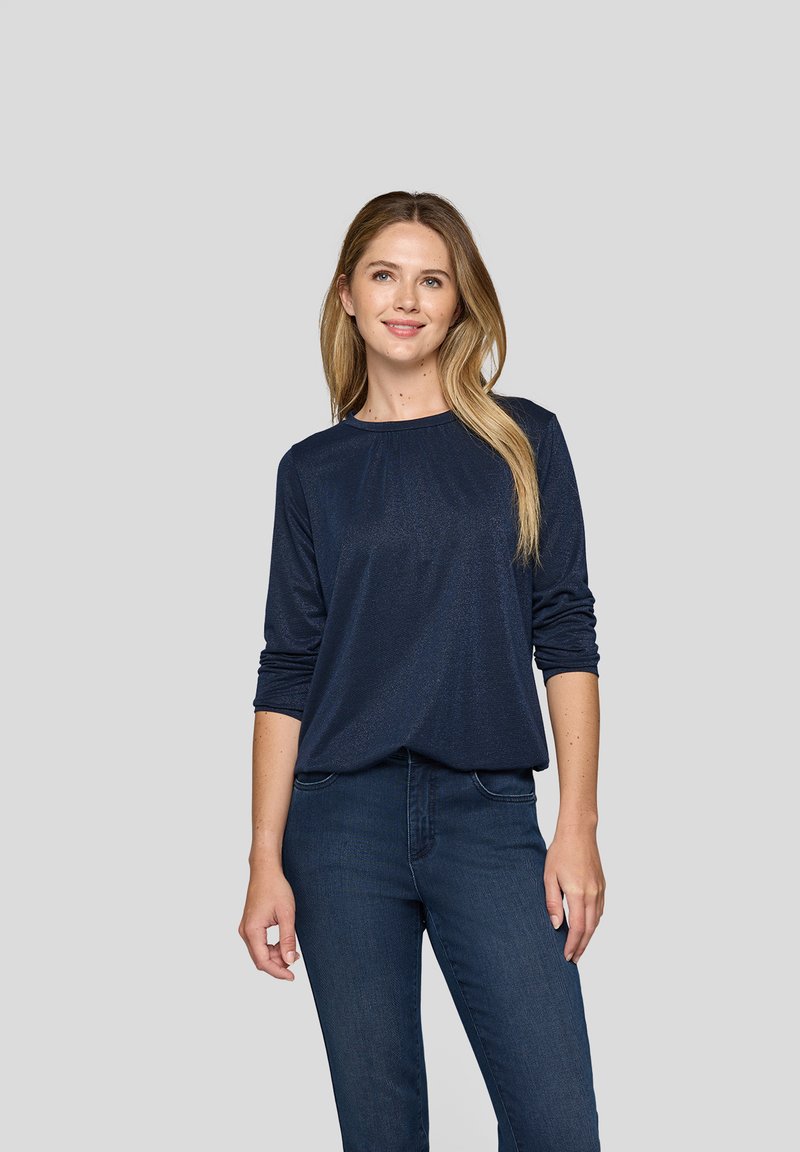 Junge Frau mit langen blonden Haaren, die ein navyblaues Langarmoberteil und dunkelblaue Jeans trägt, steht vor einem schlichten grauen Hintergrund.