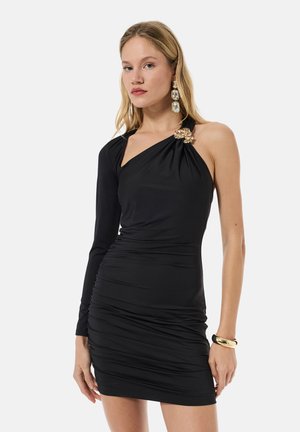 Mujer con vestido negro asimétrico de una manga, con broche dorado en forma de flor en el hombro, grandes pendientes rectangulares y pulsera dorada.